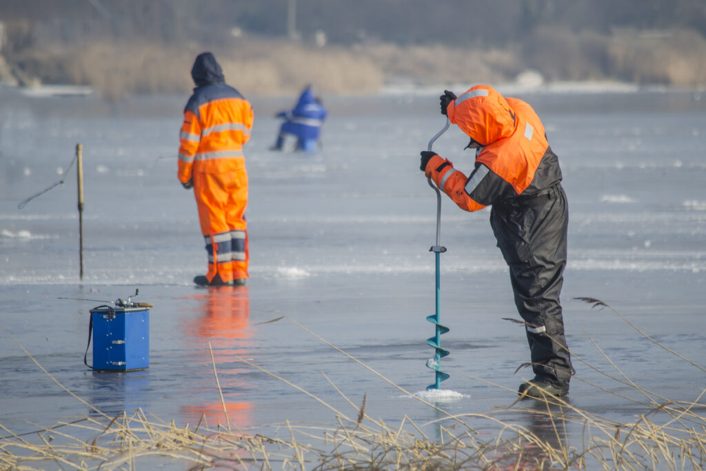 Worker drills into lake ice while others conduct winter field measurements.