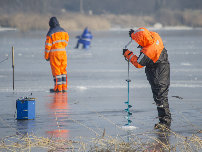Worker drills into lake ice while others conduct winter field measurements.