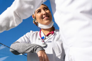 A woman smiles brightly in this waist-up shot taken in a sunny setting.