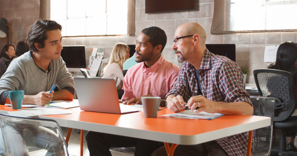 Three colleagues collaborate around a laptop at a table in a modern office workspace.