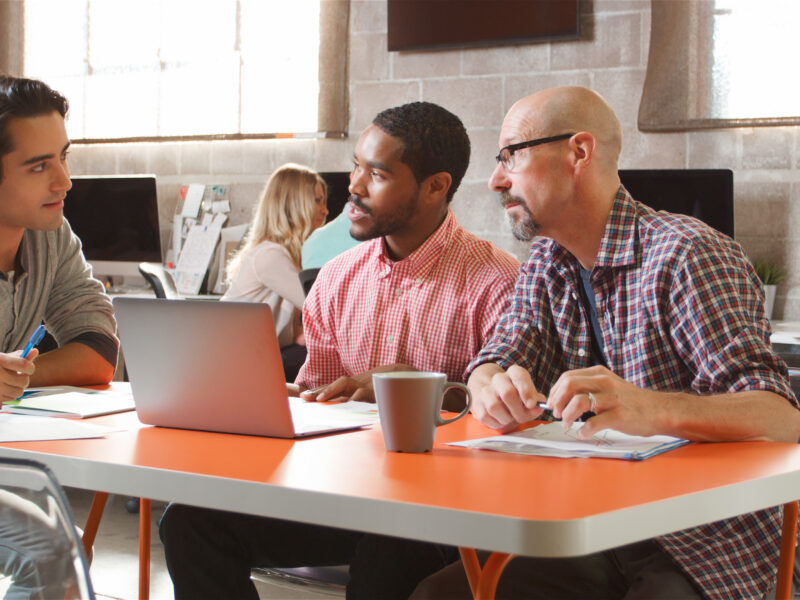 Three colleagues collaborate around a laptop at a table in a modern office workspace.
