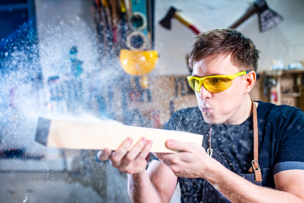 Person wearing safety glasses blowing sawdust off a wooden board in a workshop.