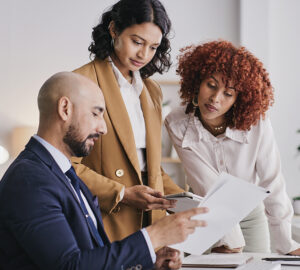 Three colleagues review paperwork together in an office, with one seated and two standing.