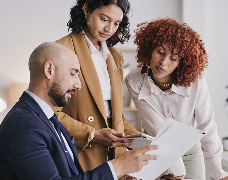Three colleagues review paperwork together in an office, with one seated and two standing.