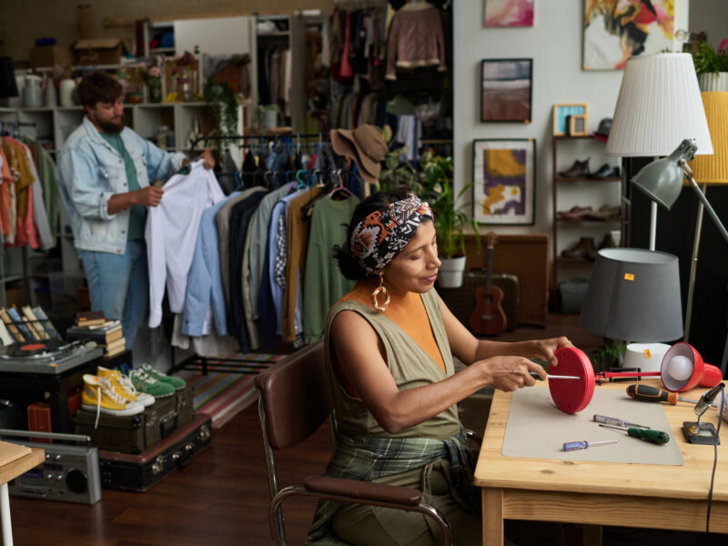 Woman fixing bottom part of secondhand electric lamp with screws while sitting against male customer choosing apparel.
