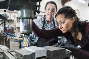 Two women wearing safety glasses standing in a metal workshop, working on metal drilling machine.