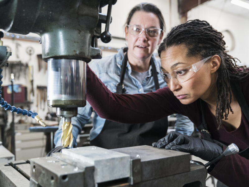 Two women wearing safety glasses standing in a metal workshop, working on metal drilling machine.